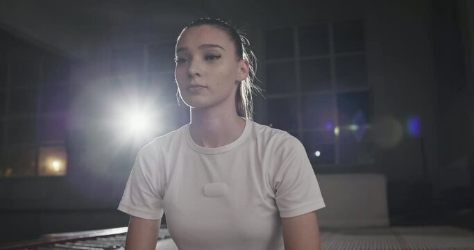 Low angle of serious confident young female gymnast sitting on trampoline while resting during acrobatic training in sports center