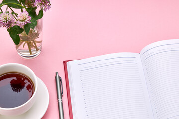 above view for pink desk with red diary of notepad and tea cup on it. table decorated with flowers. office workplace background backdrop