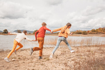 Fototapeta premium Preteen kids, boy and twin girls running in fall park with lake. Friends wearing warm knit sweaters