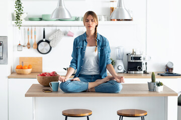 Pretty young woman in lotus position sitting on the table while relaxing in the kitchen at home.