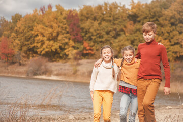 Preteen kids, boy and twin girls walking in fall park with lake.  Friends wearing warm knit sweaters