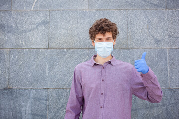 Young European man wearing safety protection medical face mask and rubber gloves, showing thumb up, looking to the camera, on gray background. Healthcare concept.