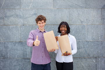 European young curly man showing thumb up and African American woman with happy face. International friends holding food bags outside on gray background. Eating and people concept.