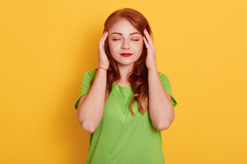 Portrait of charming, stylish woman in green shirt having head ache, touching temples with fingers and close eyes, standing over yellow background.
