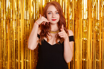 Young red haired woman wearing black elegant dress posing isolated over wall with golden tinsel on...