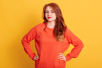 Red haired female wearing orange sweater looking directly at camera with puzzled facial expression, lady with beautiful wavy hair keeping hands son hips.