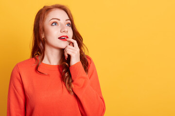 Beautiful trendy girl in orange sweater being deep in thoughts, looking in distance with finger on lips. Attractive young woman standing over yellow background.