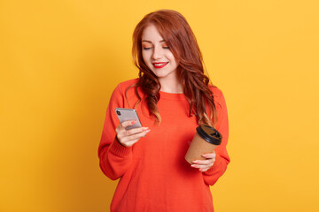 Adorable girl wearing orange sweater posing isolated over yellow background, holding coffee to take away and mobile phone, looks at device's screen with smile.