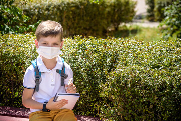 One pupil boy wearing mask is sitting on bench near his school during covid-19 pandemic time. New...