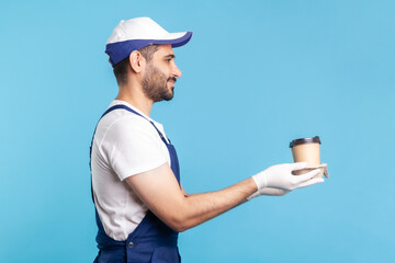 Delivery service. Side view, friendly happy courier man in overalls offering coffee, wearing safety gloves giving drinks in disposable cups and smiling. studio shot isolated on blue background