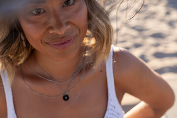 Close up portrait of a young beautiful Thai woman wearing a wooden hat and stylish necklace relaxing alone on the beach near the Atlantic Ocean. Woman's freedom lifestyle concept. Lisbon, Portugal