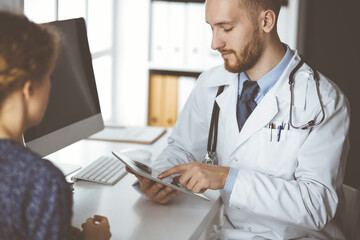 Friendly red-bearded doctor and patient woman discussing current health examination while sitting in clinic. Medicine concept