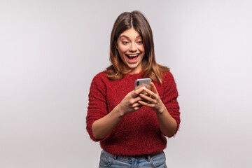Fototapeta na wymiar Amazed brunette girl in shaggy sweater using mobile phone with surprised expression, typing or reading shocking message on social network, online communication. studio shot isolated on gray background