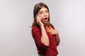 Portrait of astonished brunette woman in shaggy sweater talking with friend on cell phone, looking...