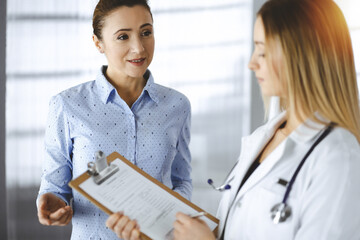 Young woman-doctor and her patient are discussing patient's current health examination, while...