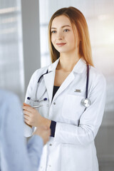 Young woman-doctor and her patient are discussing patient's current health examination, while standing together in a sunny hospital office. Female physician is writing some marks, using a clipboard