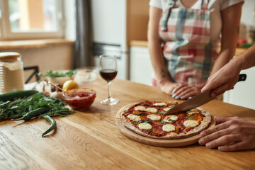 Top pizza. Cropped shot of man, cook cutting hot freshly baked pizza on the table while his girlfriend waiting. Couple making pizza together. Hobby, lifestyle