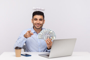 Hey you, make money online! Angelic rich businessman with nimbus on head pointing to camera and holding dollars, encouraging to earn on internet, sitting at laptop workplace. studio shot isolated
