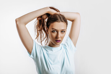 Fototapeta premium A beautiful brunette girl holds her hair and makes her own hairstyle. Young model with long hair, fashion makeup with purple lips on an gray background. The woman seriously looking at the camera.