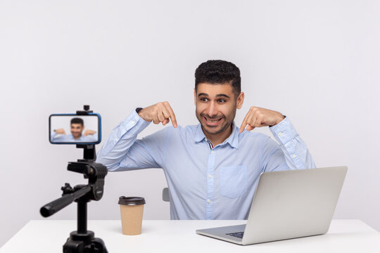 Online webinar, distant training. Successful businessman sitting at workplace, pointing down subscribe gesture and smiling excitedly to mobile phone while recording course, doing blog. studio shot