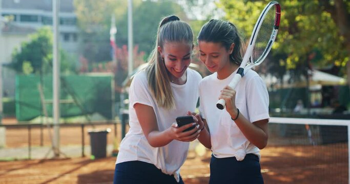 Authentic shot of young female teenage tennis players friends are having fun using a smartphone after a friendly match or training workout game on the court in a sunny day.