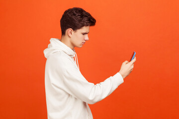 Profile portrait of young concentrated man in casual hoodie looking at screen of mobile phone,...