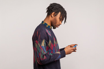 Profile portrait of african man with dreadlocks concentrated looking at smartphone display, absorbed with virtual life, internet connection. Indoor studio shot isolated on gray background