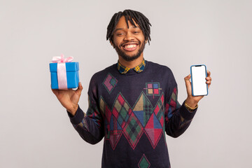Cheerful pleased african guy with dreadlocks holding empty blank smartphone and gift box in hands with toothy smile on face, online shopping, bonuses. Indoor studio shot isolated on gray background
