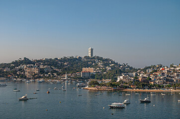 Acapulco, Mexico - November 25, 2008: Pharos of Santa Lucia behind yacht harbor with hill and tall building in back under twilight sky. Blue bay water in front of boardwalk with housing.