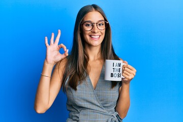 Young hispanic woman drinking from i am the boss coffee cup doing ok sign with fingers, smiling...