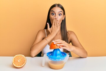 Young hispanic woman sitting on the table using juicer covering mouth with hand, shocked and afraid...