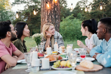 Young joyful couples eating and talking by served festive table under pine tree