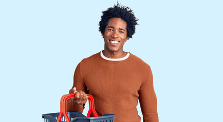 Handsome african american man with afro hair holding supermarket shopping basket looking positive and happy standing and smiling with a confident smile showing teeth