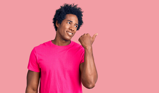Handsome african american man with afro hair wearing casual pink tshirt smiling with happy face looking and pointing to the side with thumb up.