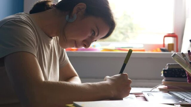 concentrated young brunette woman designer draws in sketchbook with marker sitting at table with laptop near window in room closeup