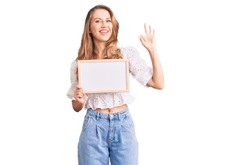 Fototapeta premium Young caucasian woman with blond hair holding blank empty banner doing ok sign with fingers, smiling friendly gesturing excellent symbol