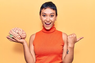 Young woman holding brain pointing thumb up to the side smiling happy with open mouth