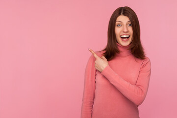 Obraz premium Happy surprised woman in pink sweater pointing finger away with toothy smile, wondering about empty space for your advertisement, template. Indoor studio shot isolated on pink background