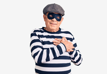 Senior handsome man wearing burglar mask and t-shirt smiling with hands on chest with closed eyes and grateful gesture on face. health concept.