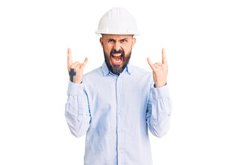 Young handsome man wearing architect hardhat shouting with crazy expression doing rock symbol with hands up. music star. heavy concept.