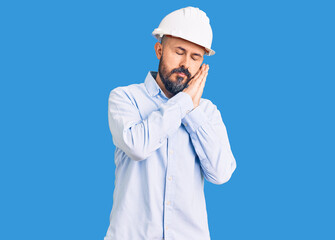 Young handsome man wearing architect hardhat sleeping tired dreaming and posing with hands together while smiling with closed eyes.
