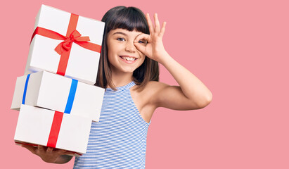 Young little girl with bang holding gifts smiling happy doing ok sign with hand on eye looking through fingers