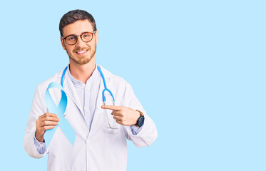 Handsome young man with bear wearing doctor uniform and holding blue ribbon smiling happy pointing with hand and finger