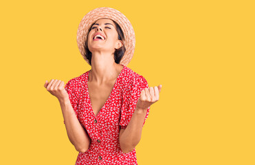 Young beautiful woman wearing summer hat very happy and excited doing winner gesture with arms raised, smiling and screaming for success. celebration concept.