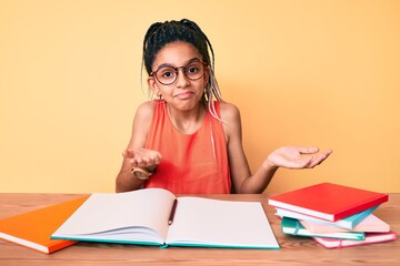 Young african american girl child with braids studying for school exam clueless and confused expression with arms and hands raised. doubt concept.