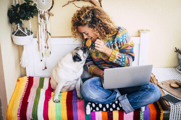 Cheerful beautiful young woman share a cookie with her funny and lovely pug dog while work with laptop computer at home on a coloured cover - concept of love for animals and modern people