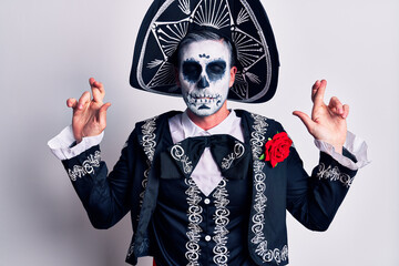 Young man wearing mexican day of the dead costume over white gesturing finger crossed smiling with hope and eyes closed. luck and superstitious concept.