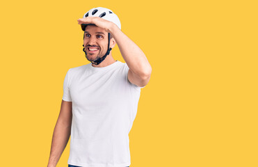 Young handsome man wearing bike helmet very happy and smiling looking far away with hand over head. searching concept.