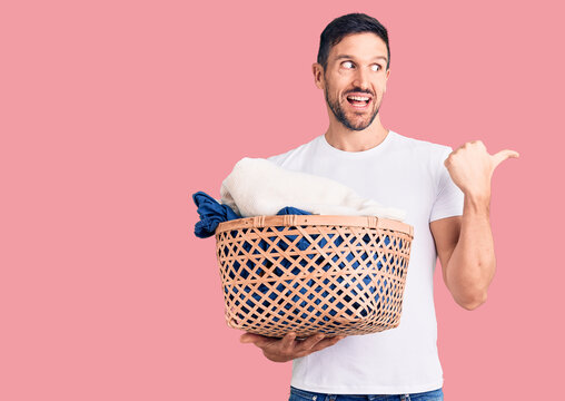 Young handsome man holding laundry basket pointing thumb up to the side smiling happy with open mouth