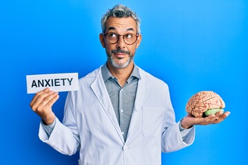 Middle age grey-haired man wearing doctor coat holding brain and anxiety message smiling looking to...